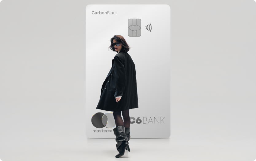 Mulher de preto caminha à frente de um Cartão C6 Carbon Mastercard Black prata em tamanho ampliado, sobre fundo claro.
