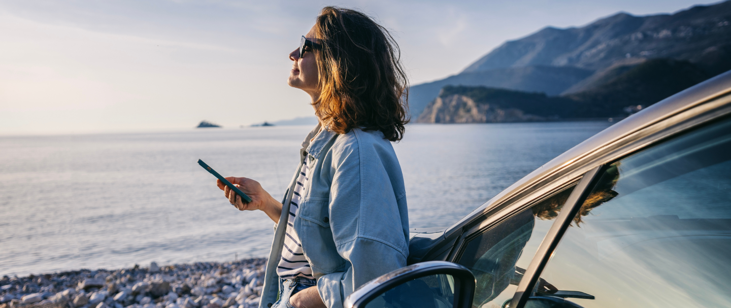 Mulher apoiada em carro Fotografia de mulher de cabelo curto, ondulado e castanho, usando óculos escuro, apoiada na lateral de um carro, segurando um celular com a mão direita e observando a paisagem de lago e montanhas.