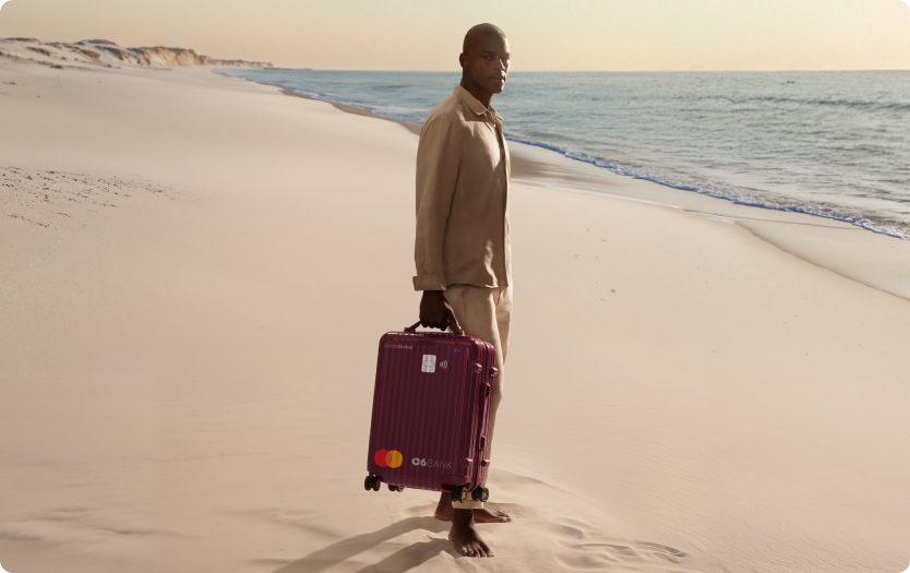 Homem na praia puxa mala com layout do C6 Conta Global. Cena ao entardecer transmite mobilidade e tranquilidade.