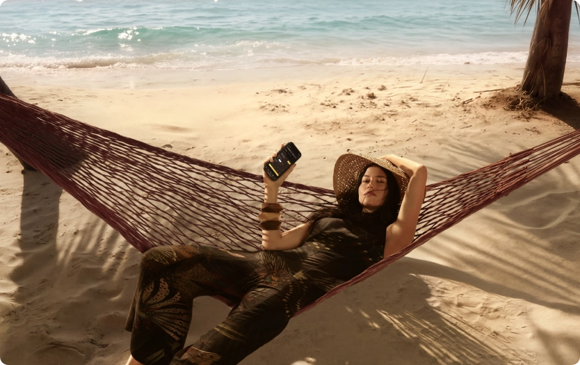 Mulher deitada em rede sobre a areia com mar ao fundo, usando chapéu e roupa estampada, segurando celular com um braço apoiado atrás da cabeça.