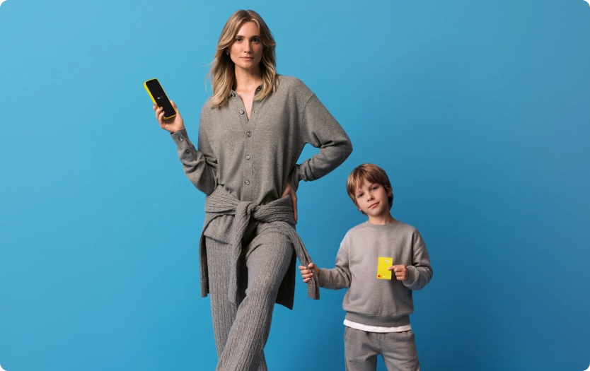 Mãe e filho em fundo azul. Mulher adulta segurando celular exibindo o app C6 Bank e com bola de futebol apoiada em um dos pés. Ao lado, criança segurando cartão C6 Yellow na cor amarela,