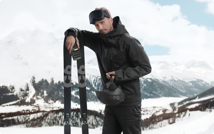 Homem em paisagem de neve com montanhas ao fundo, usando roupa de inverno, apoiada em esquis, segurando capacete, com celular exibindo app do C6 Bank, posicionado sobre o equipamento.