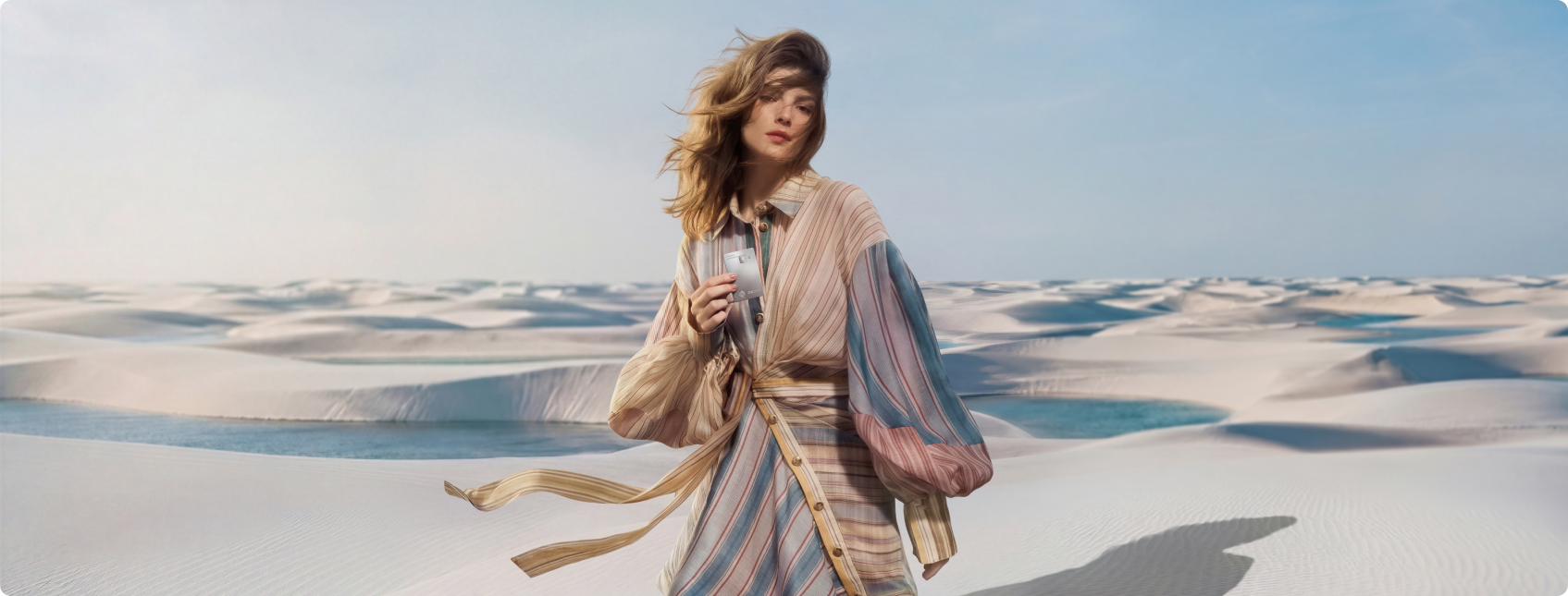 Mulher em paisagem de dunas de areia, usando roupa listrada e segurando cartão C6 Graphene junto ao corpo, com tecido sendo movimentado pelo vento.