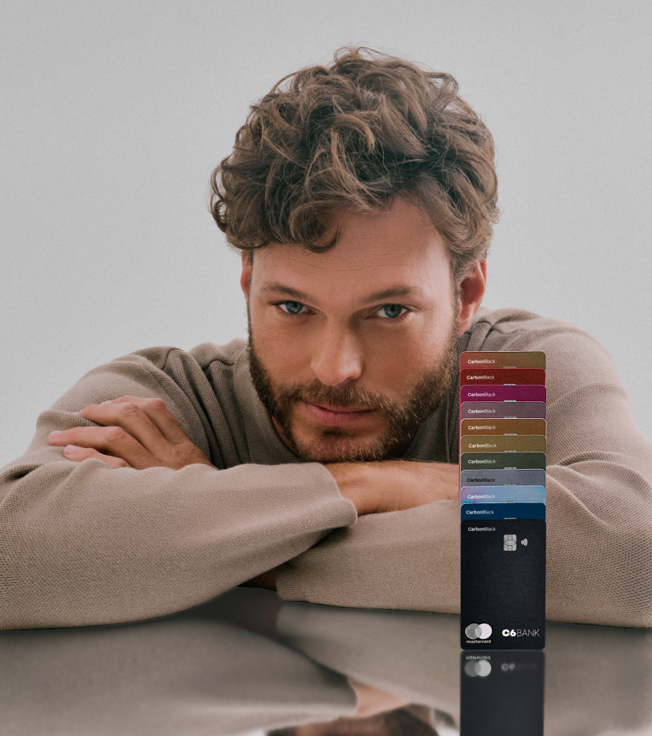 Foto de um homem branco de cabelos e barba castanhos claro, onde ele apoia seus braços em uma mesa refletiva com múltiplos cartões C6 Bank de diversas cores empilhados um em cima do outro ao seu lado. Ele veste um suéter bege, tem olho claro e tem uma expressão facial confiante.