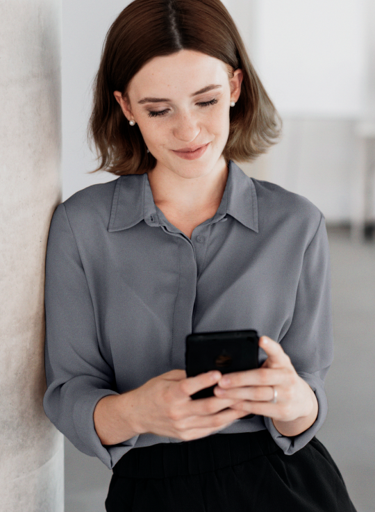 Mulher de camisa cinza encostada em uma parede, segurando um celular preto e olhando para ele com um leve sorriso. Ambiente de escritório ao fundo.