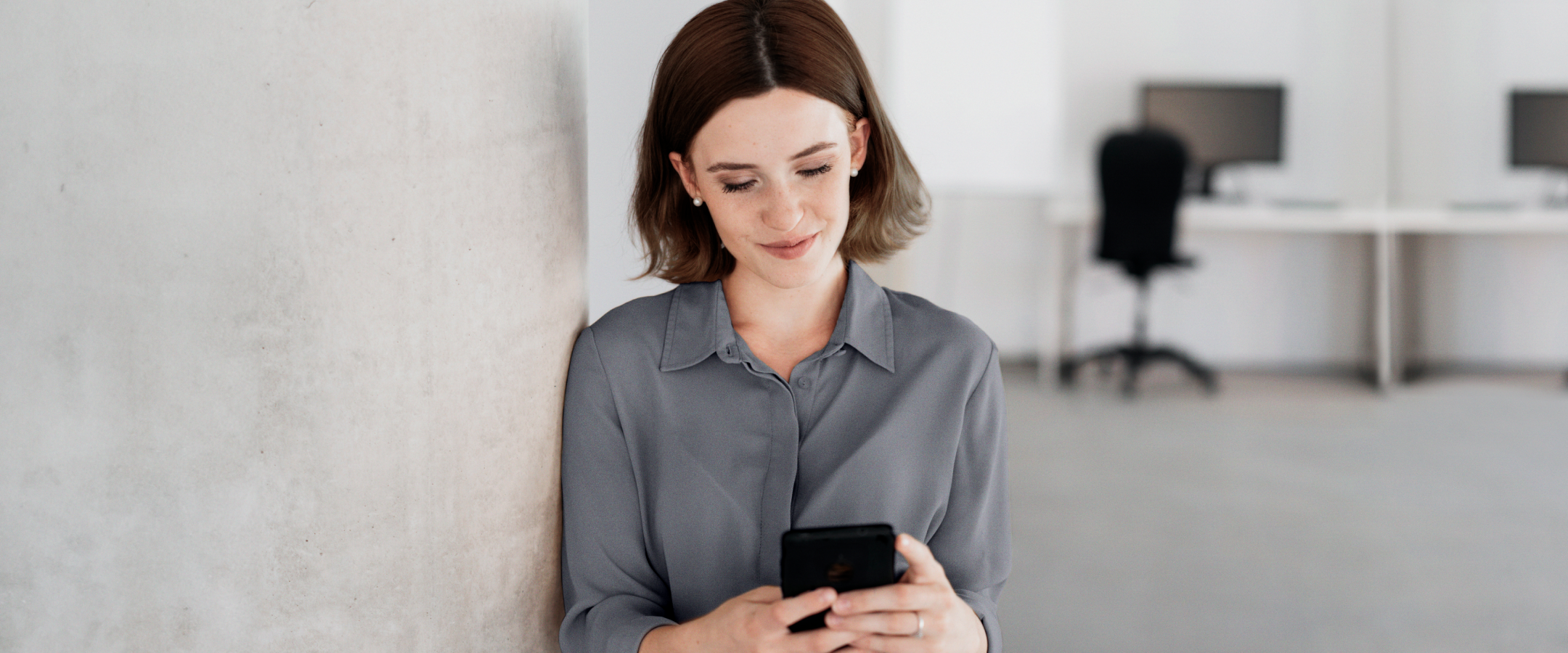 Mulher de camisa cinza encostada em uma parede, segurando um celular preto e olhando para ele com um leve sorriso. Ambiente de escritório ao fundo.