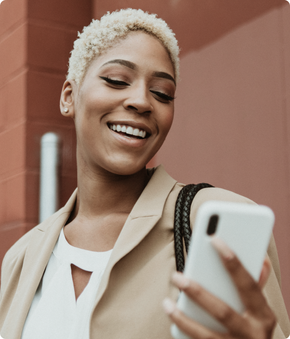 Mulher sorridente com cabelo loiro curto segura um celular branco e olha para a tela. Fundo com parede vermelha em ambiente externo.