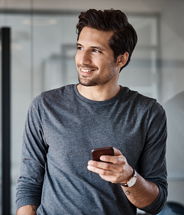 Homem jovem, sorrindo, segura um celular com a mão direita e olha para o lado, em um ambiente interno com paredes de vidro.