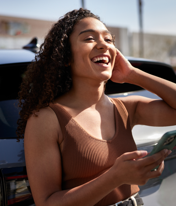 Mulher jovem de cabelos cacheados, sorrindo, segura um celular verde e encosta-se na traseira de um carro, em um ambiente externo ensolarado.