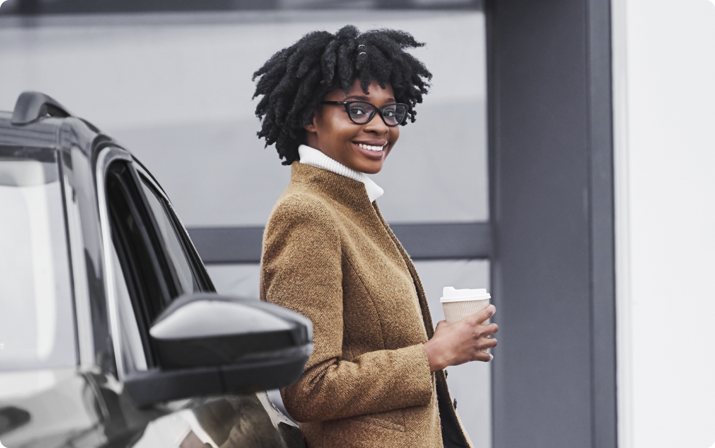 Mulher negra de pé, encostada em um carro preto, segurando um copo de café na sua mão direita e vestindo um casaco bege.