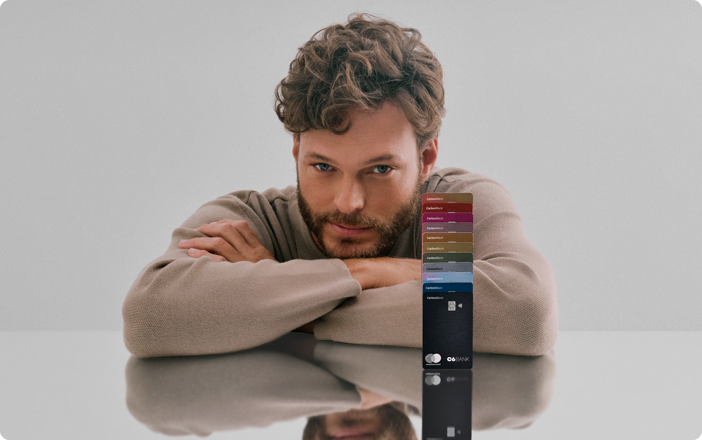 Foto de um homem branco de cabelos e barba castanhos claro, onde ele apoia seus braços em uma mesa refletiva com múltiplos cartões C6 Bank de diversas cores empilhados um em cima do outro ao seu lado. Ele veste um suéter bege, tem olho claro e tem uma expressão facial confiante.