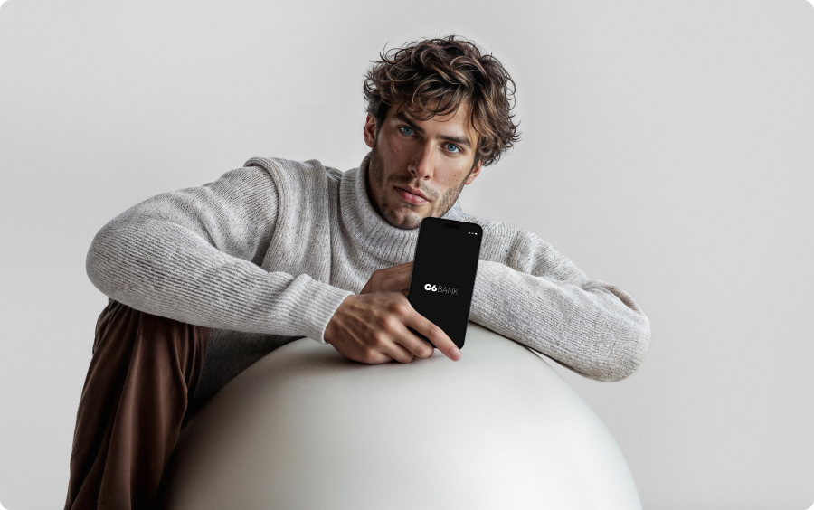 Homem branco de cabelo castanho e olhos claros, sentado no chão, apoiado sobre uma bola de yoga na cor cinza, segurando em sua mão direita um celular aberto com o logo do C6 Bank na tela.