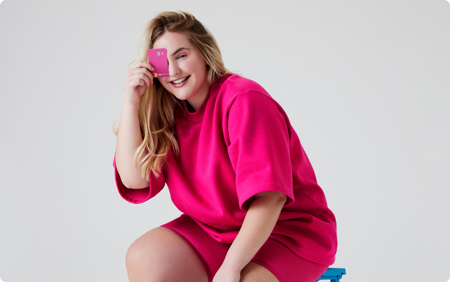 Mulher branco, com cabelo loiro, vestindo um conjunto de camiseta e shorts na cor pink, segurando o cartão Pink da C6 Yellow em frente ao seu olho direito.