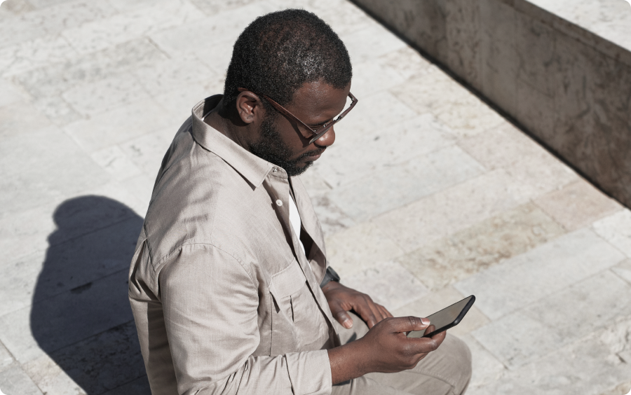 Homem negro sentado em um degrau, vestindo uma roupa bege, segurando um celular em sua mão direita.