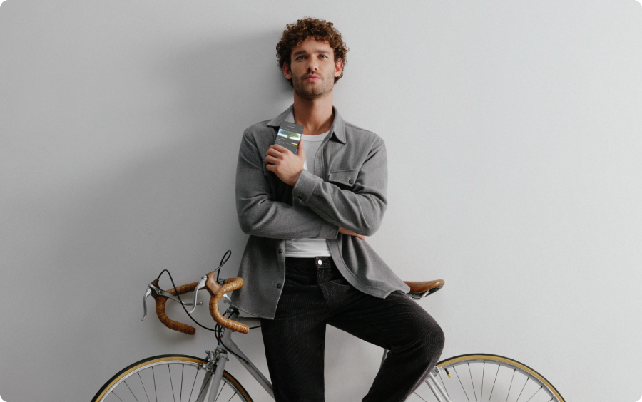 Foto de um homem branco, cabelo castanho encaracolado, apoiando seu corpo em uma bicicleta encostada em uma parede. Ele segura próximo ao peito um celular na tela do aplicativo C6 Bank mostrando o extrato de carbono. Ele veste um blazer cinza por cima de uma camiseta branca e uma calça preta.