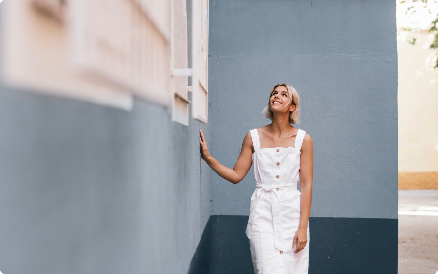 Mulher branca e loira, usando um vestido midi branco, de pé, encostando a mão em uma parede azul, com janelas brancas.