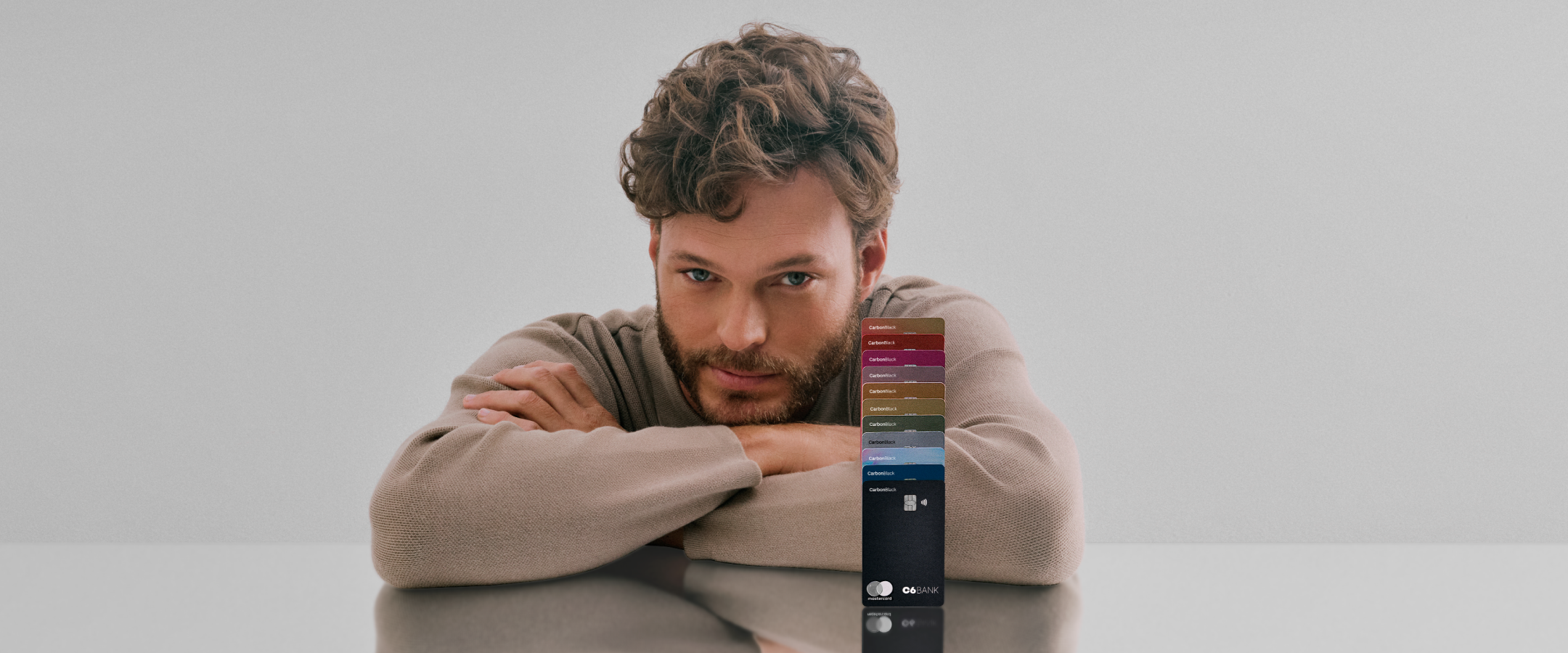 Foto de um homem branco de cabelos e barba castanhos claro, onde ele apoia seus braços em uma mesa refletiva com múltiplos cartões C6 Bank de diversas cores empilhados um em cima do outro ao seu lado. Ele veste um suéter bege, tem olho claro e tem uma expressão facial confiante.