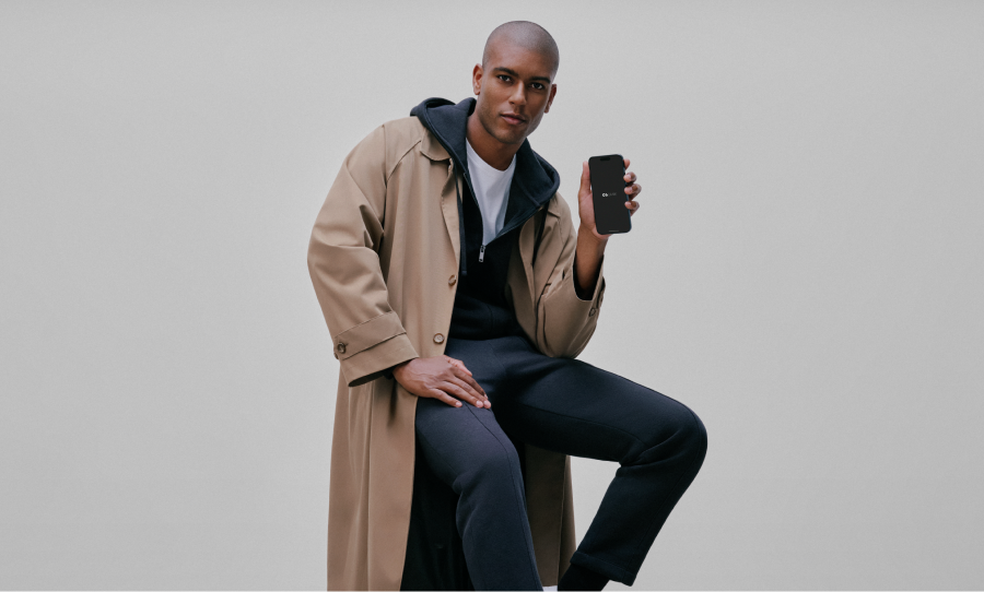 Foto de um homem confiante de pele preta que tem seu cabelo raspado. Ele senta em um banco segurando um celular com a tela preta e o logo do C6 Bank no centro. Veste um sobretudo bege por cima de uma blusa azul marinho e uma camiseta branca.