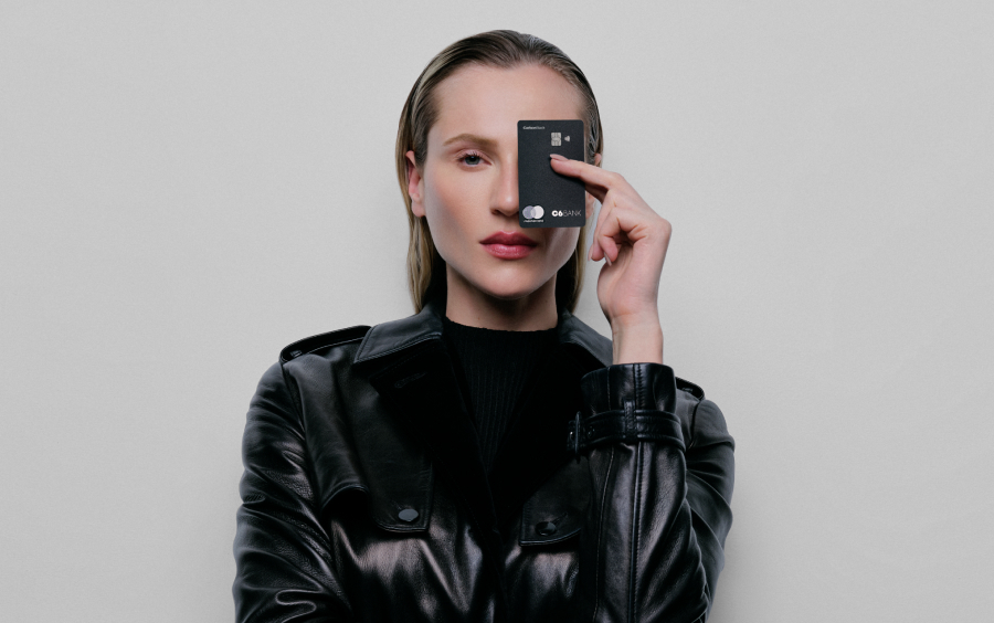 Foto de mulher branca de cabelos loiros e olhos claros. Veste um sobretudo de couro preto e segura um cartão C6 Bank preto com a mão esquerda cobrindo seu olho esquerdo. Tem expressões faciais confiantes.