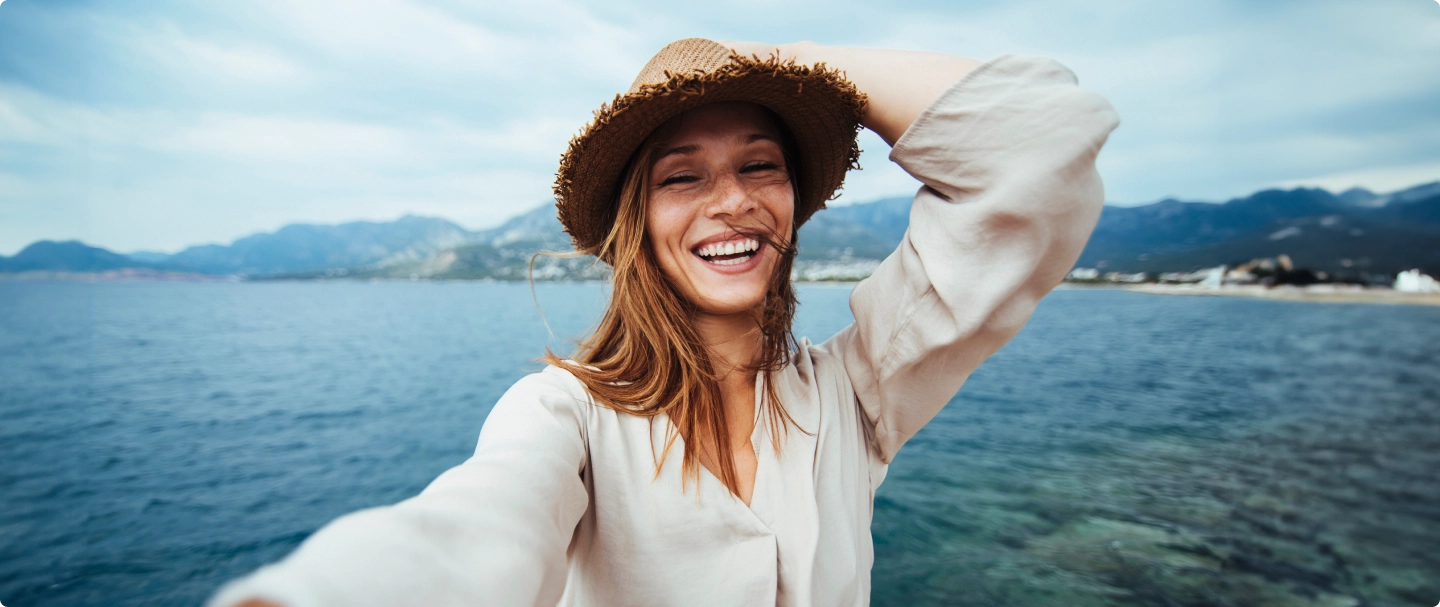 Selfie de mulher loira, segurando um chapéu de palha com a mão esquerda em um fundo com um mar e uma montanha
