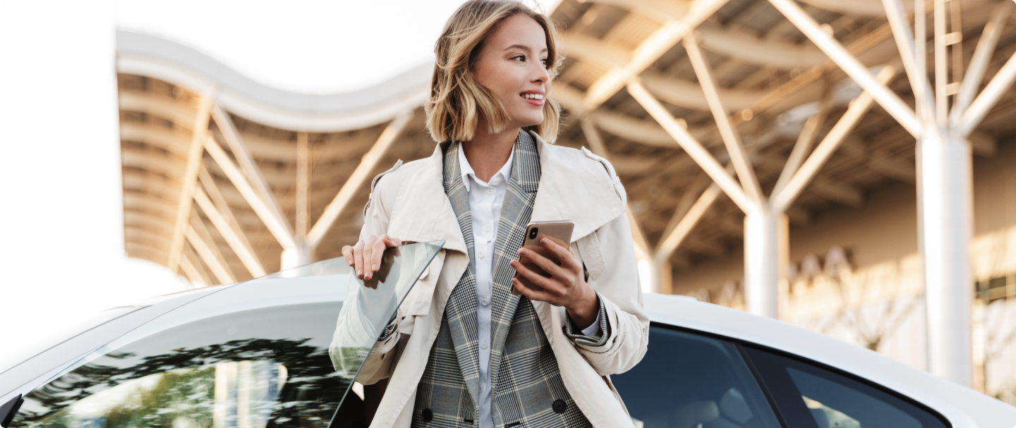 Fotografia de mulher loira usando um trenchcoat com blazer xadrez embaixo, encostada em um carro branco, segurando um celular em sua mão esquerda