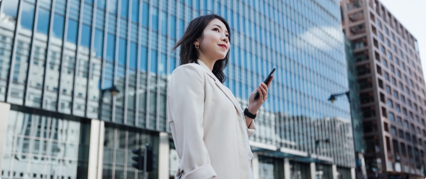Foto de mulher asiática de terno branco segurando um celular em sua mão em frente a um prédio espelhado
