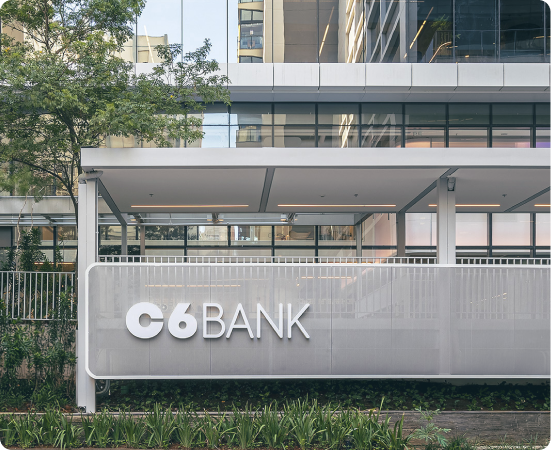 Fachada do C6 Bank com logo em painel branco, estrutura moderna de vidro e metal, cercada por vegetação.