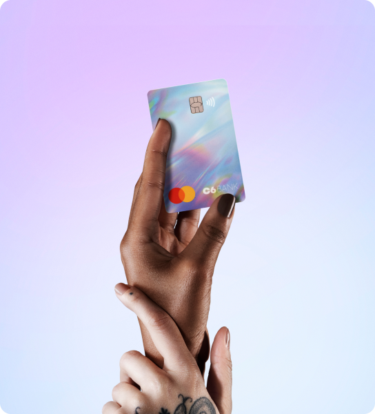 Cartão Rainbow do C6 Bank com holográfico iridescente, segurado por duas mãos contra fundo lilás degradê.