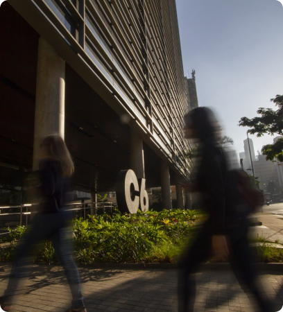 Fachada do C6 Bank com logotipo e vegetação, em dia ensolarado, com pessoas caminhando em frente ao prédio.