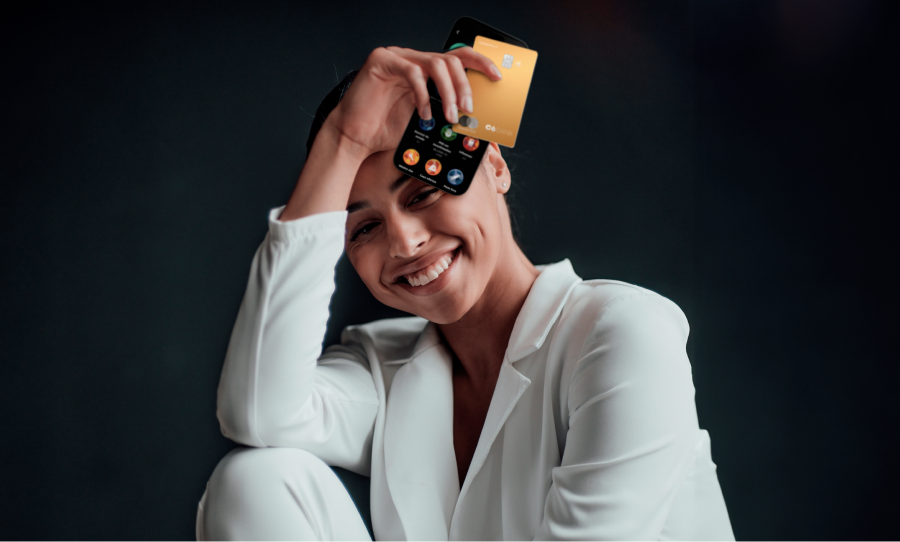 Foto de mulher vestindo blazer e calça branca, segurando com sua mão direita, acima de seu olho esquerdo, um celular com a tela de C6 Experience e um cartão dourado.