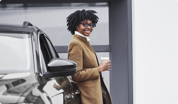 Mulher negra de pé, encostada em um carro preto, segurando um copo de café na sua mão direita e vestindo um casaco bege.