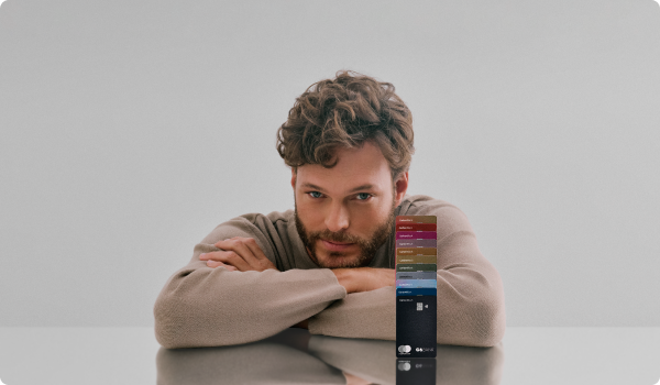 Foto de um homem branco de cabelos e barba castanhos claro, onde ele apoia seus braços em uma mesa refletiva com múltiplos cartões C6 Bank de diversas cores empilhados um em cima do outro ao seu lado. Ele veste um suéter bege, tem olho claro e tem uma expressão facial confiante.