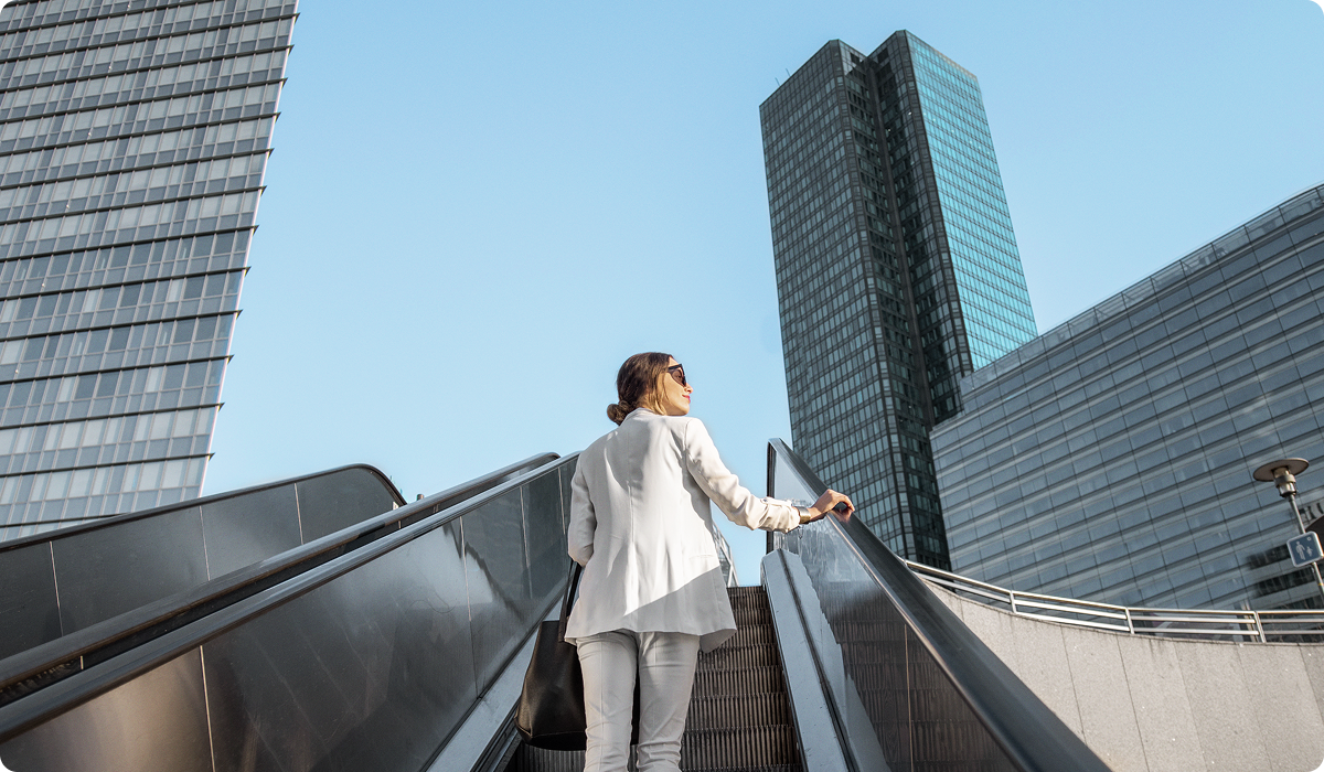 Mulher de cabelos presos e óculos escuros, vestindo blazer e calça brancos, sobe uma escada rolante ao ar livre. Ela carrega uma bolsa preta no ombro e olha para cima, com edifícios altos e modernos ao fundo. O céu está claro e sem nuvens, sugerindo um ambiente urbano corporativo.