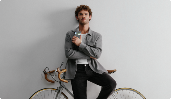 Foto de um homem branco, cabelo castanho encaracolado, apoiando seu corpo em uma bicicleta encostada em uma parede. Ele segura próximo ao peito um celular na tela do aplicativo C6 Bank mostrando o extrato de carbono. Ele veste um blazer cinza por cima de uma camiseta branca e uma calça preta.