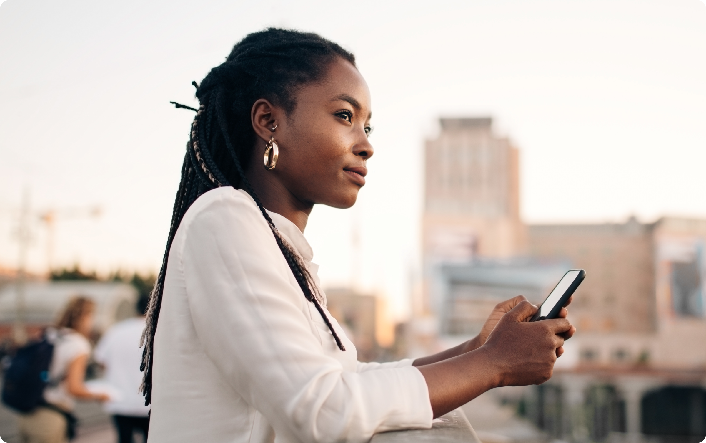 Mulher negra de pé, encostada em um parapeito, olhando para a paisagem e segurando em suas mãos um celular.