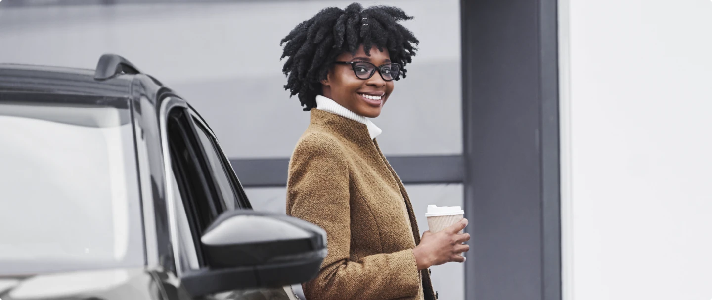 Fotografia de mulher negra vestindo um sobretudo amarelo, segurando um copo de café com a mão direta e apoiada em um carro preto, olhando e sorrindo para a câmera