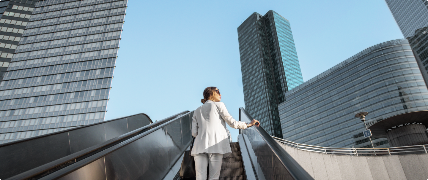 Mulher de cabelos presos e óculos escuros, vestindo blazer e calça brancos, sobe uma escada rolante ao ar livre. Ela carrega uma bolsa preta no ombro e olha para cima, com edifícios altos e modernos ao fundo. O céu está claro e sem nuvens, sugerindo um ambiente urbano corporativo.