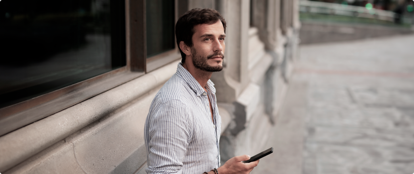 Homem branco de camisa social, apoiado na parede, segurando um celular em sua mão