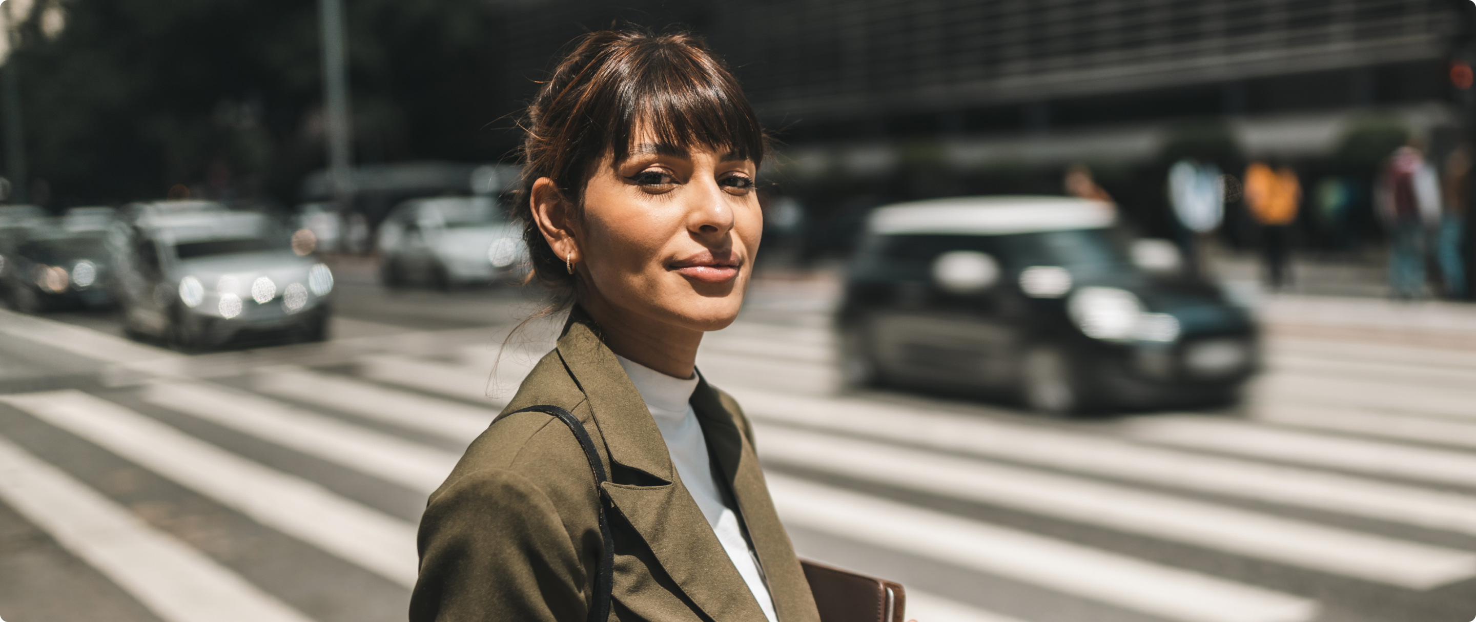 Mulher de blazer verde musgo, parada com um fundo de uma avenida, segurando um copo de café na mão e olhando para a câmera