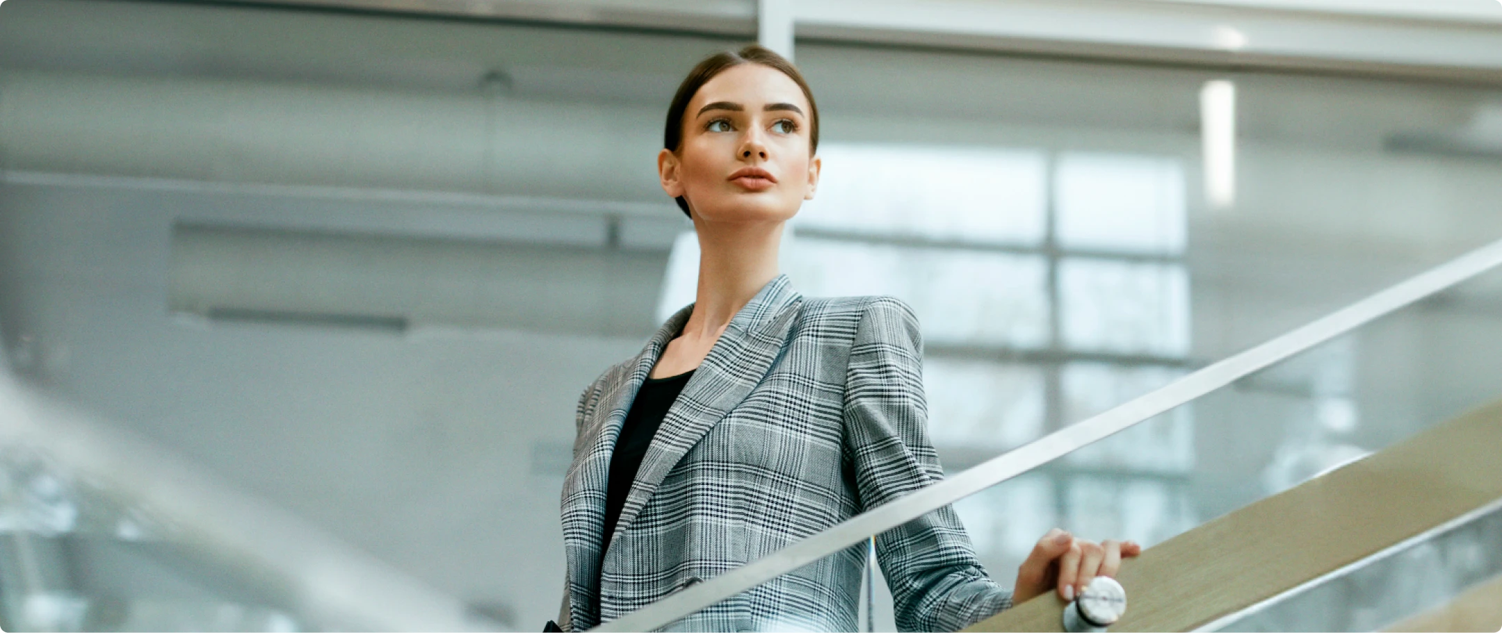 Mulher de blazer cinza e cabelo preso, descendo as escadas e segurando no corrimão.