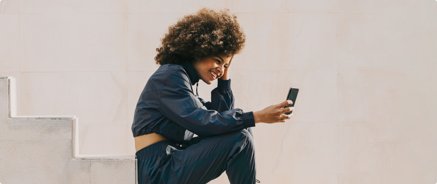 Foto de mulher parda com cabelo castanho com luzes sentada em uma escada segurando o celular com a mão direita