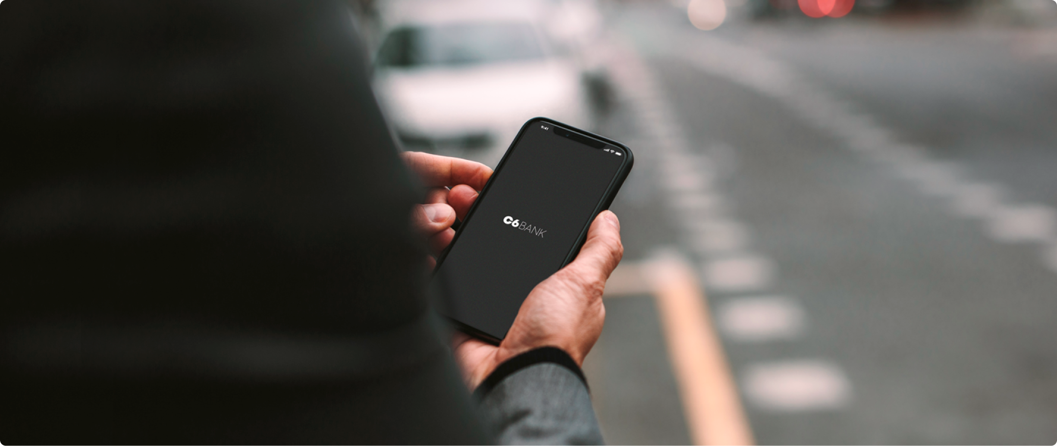 Fotografia de mão masculina segurando um celular com o logo do C6 Bank com a mão direita