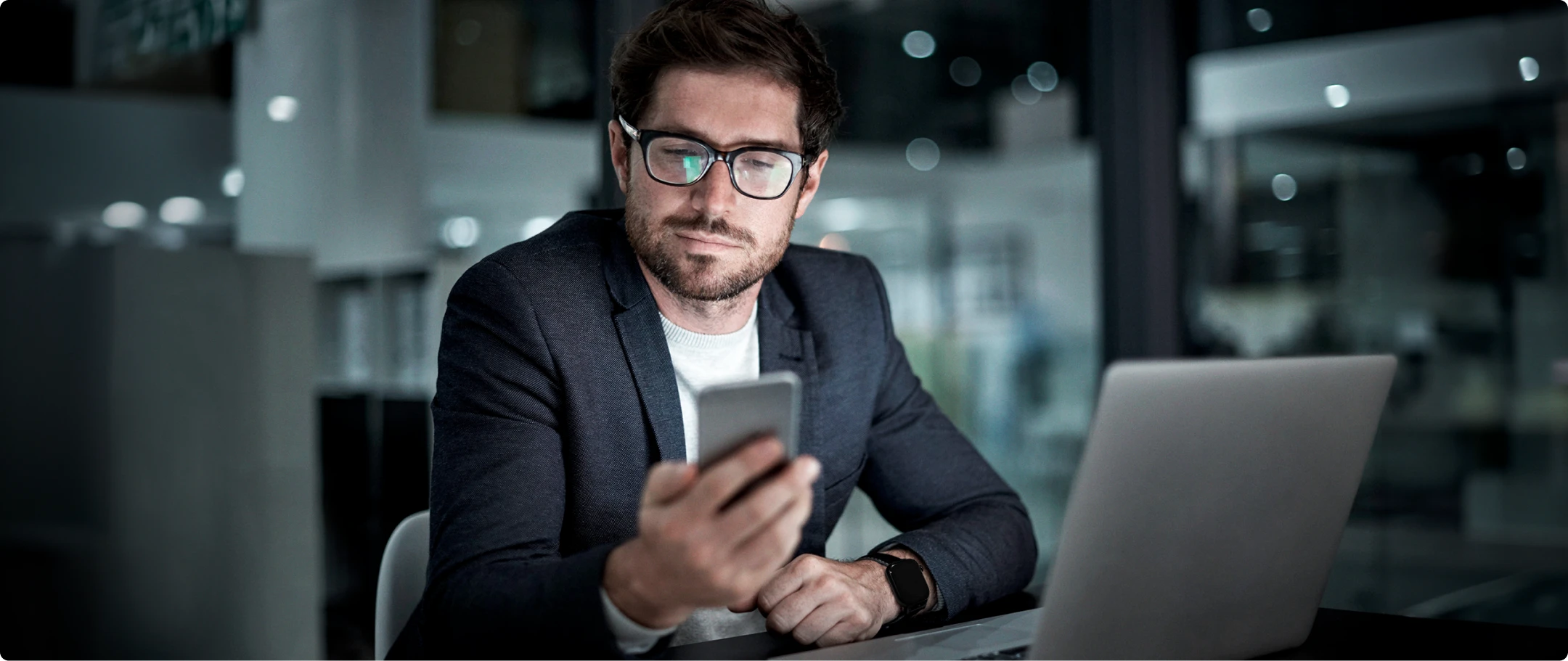 Homem de blazer azul marinho e camiseta branca segurando um celular em sua mão, sentado em frente a um computador