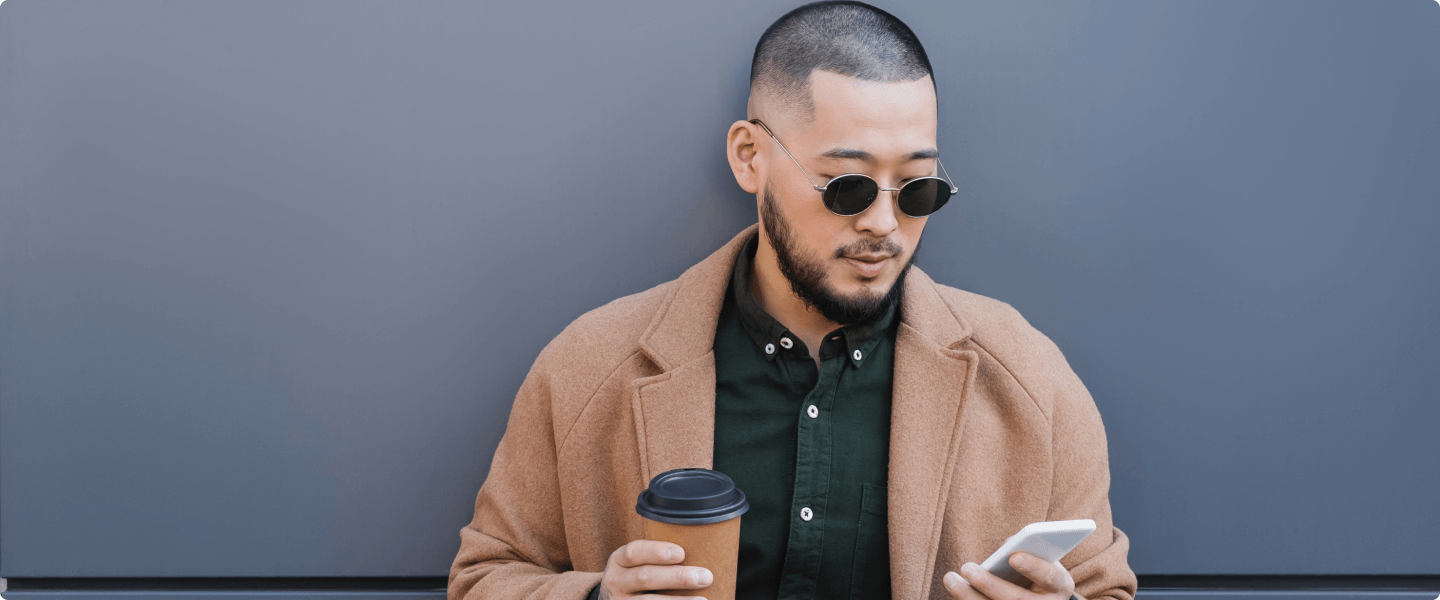 Foto de homem asiático de óculos escuros. O homem está encostado numa parede cinza onde segura um café na mão direita e um celular na mão esquerda