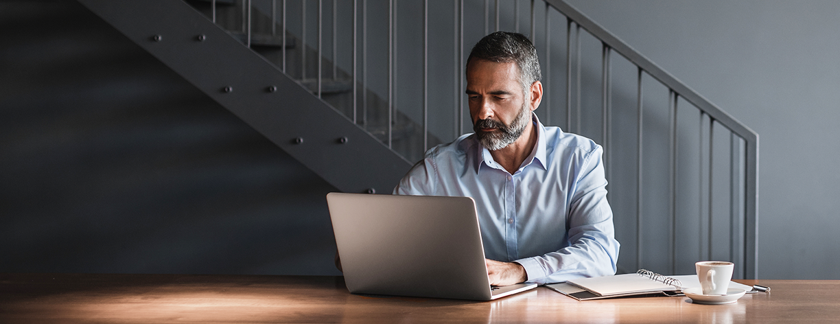 Homem grisalho vestindo camisa azul trabalha no notebook, com caderno e café ao lado, em ambiente elegante.