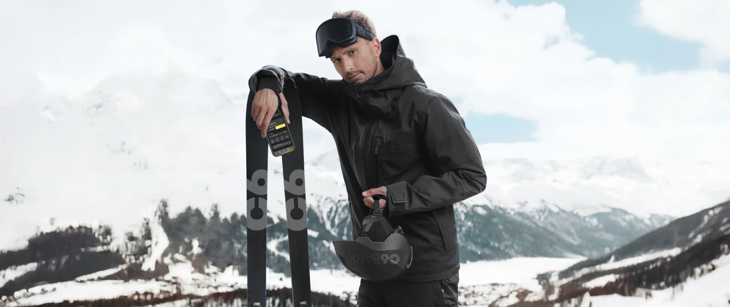 Homem com esquis na neve S 26-27 Homem em paisagem de neve com montanhas ao fundo, usando roupa de inverno, apoiada em esquis, segurando capacete, com celular exibindo app do C6 Bank, posicionado sobre o equipamento.