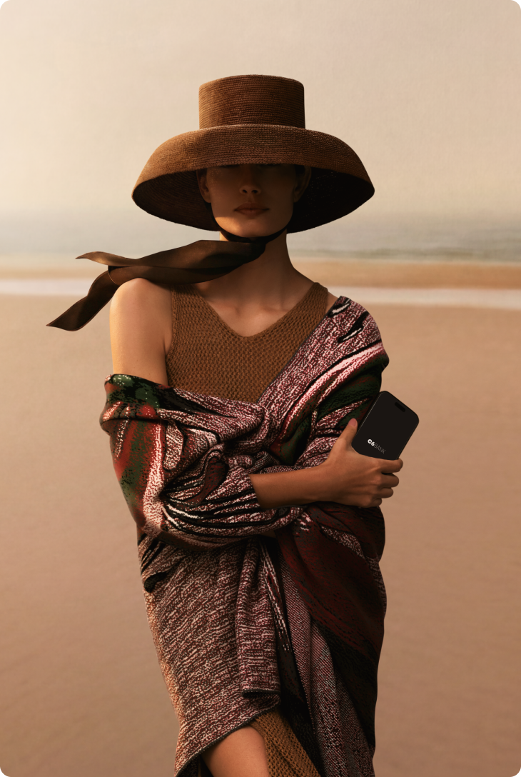 Mulher em pé na praia com mar ao fundo, usando chapéu de abas largas e roupa colorida, segurando celular junto ao corpo.