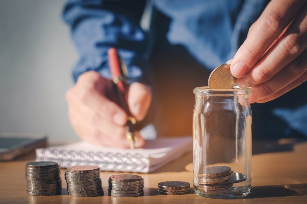 Imagem mostra as mãos de uma pessoa escrevendo em um caderno enquanto coloca moedas dentro de um pote de vidro, calculando o cadastro positivo