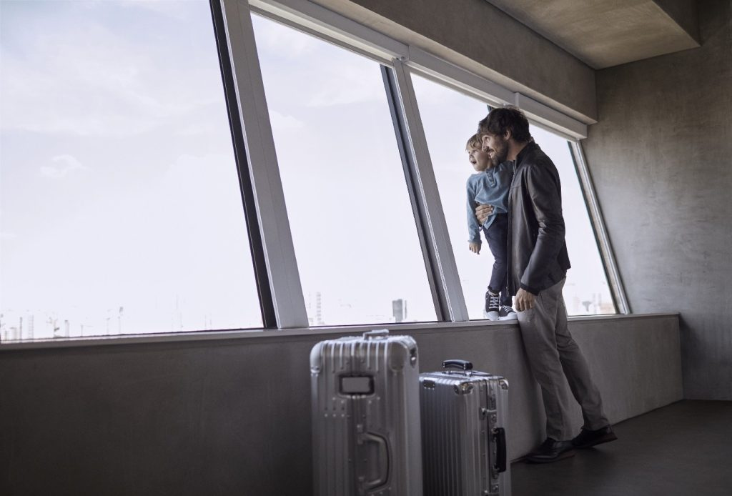 A imagem mostra um homem e uma criança com malas de viagem olhando pela janela de um aeroporto