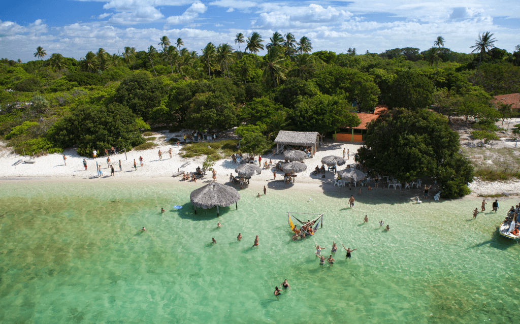 Praia de Jericoacoara, Brasil, com águas rasas e cristalinas em um dia de sol. Turistas aproveitam o cenário tropical, com vegetação nativa e cabanas de palha na areia branca.